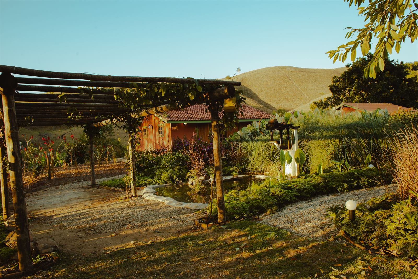 Pergolado de madeira e espelho d'água diante da sede da Fazenda Santa Helena, com os morros do Vale do Paraíba ao fundo